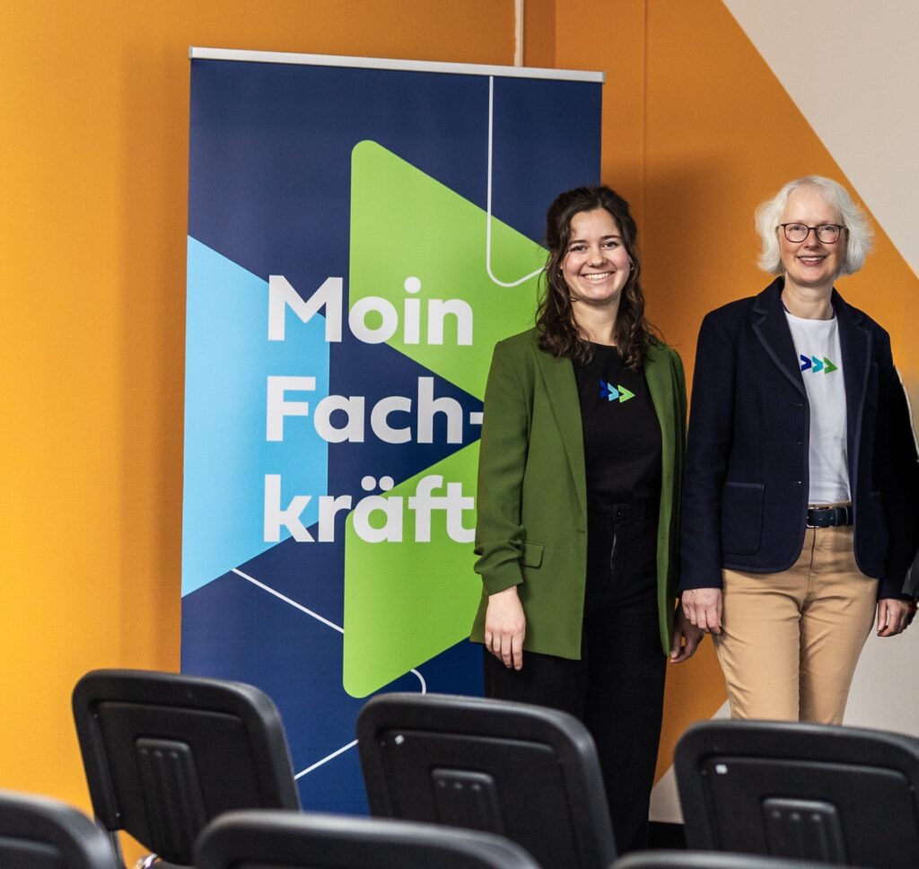 Zwei Personen stehen lächelnd vor einem „Moin Fachkräfte“-Banner in einem Seminar- oder Veranstaltungsraum.